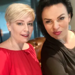 Portret dwóch uśmiechniętych kobiet z Bydgoszczy, jedna w czerwonej bluzce z odkrytymi ramionami, druga w czarnej, pozujących w biurze.