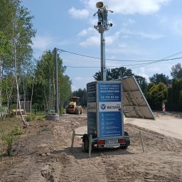Grupa Oktagon Sp. z o.o. - Mobilna wieża monitoringu Oktagon z kamerami, głośnikami i panelami słonecznymi, stojąca na piaszczystym placu budowy z widocznym sprzętem ciężkim i drzewami w tle.