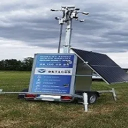 Mobilna wieża monitoringu z panelem solarnym na przyczepie, ustawiona na trawiastym terenie pod pochmurnym niebem. Widoczne kamery i oświetlenie na szczycie wieży.