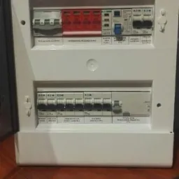 Otwarta, biała skrzynka elektryczna z widocznymi bezpiecznikami i wyłącznikami, umieszczona na drewnianej powierzchni. Widoczne napisy i oznaczenia na elementach.