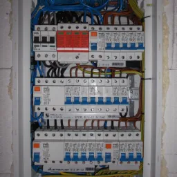 Rozdzielnica elektryczna z zainstalowanymi bezpiecznikami, ogranicznikami przepięć i przewodami w kolorach niebieskim, brązowym i żółto-zielonym, widok z bliska na białą obudowę i elementy...