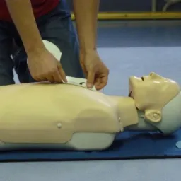 Osoba w dżinsach ćwiczy na manekinie resuscytację, przyklejając elektrody defibrylatora.