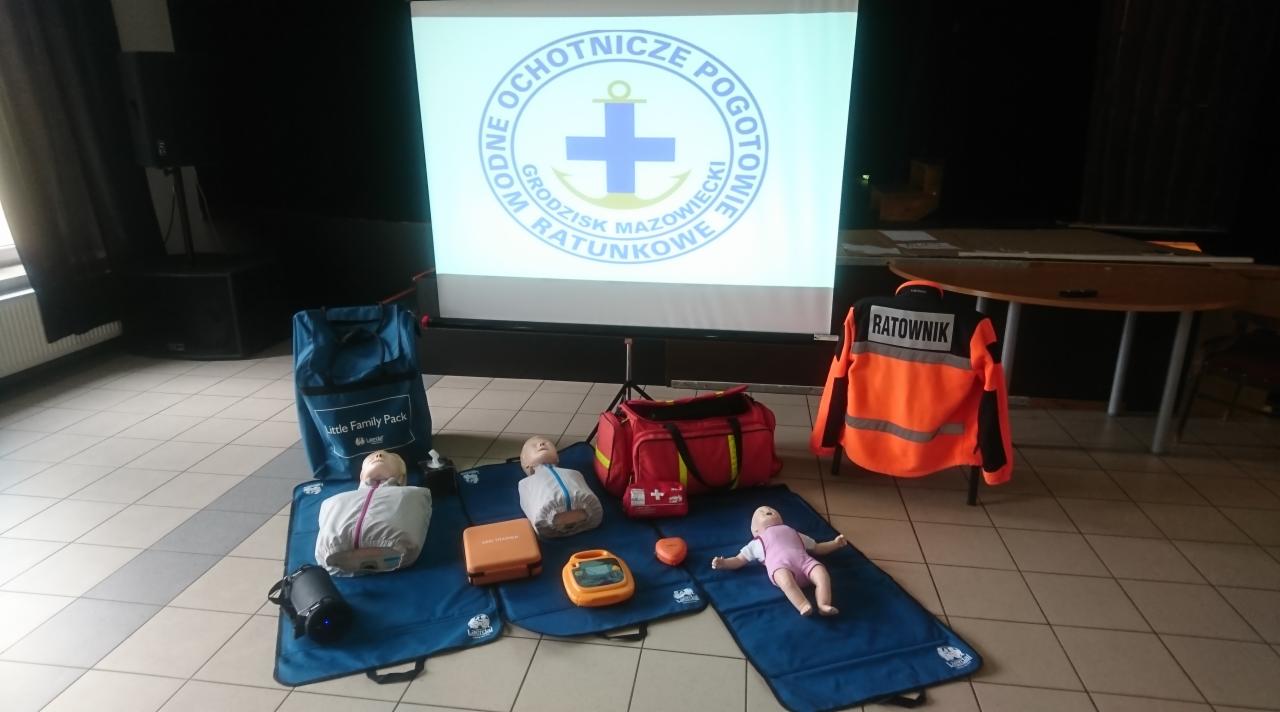 Sala szkoleniowa w Baranowie z manekinami do nauki resuscytacji, torbą ratowniczą i defibrylatorem, na ekranie wyświetlone logo Wodnego Ochotniczego Pogotowia Ratunkowego Grodzisk Mazowiecki.