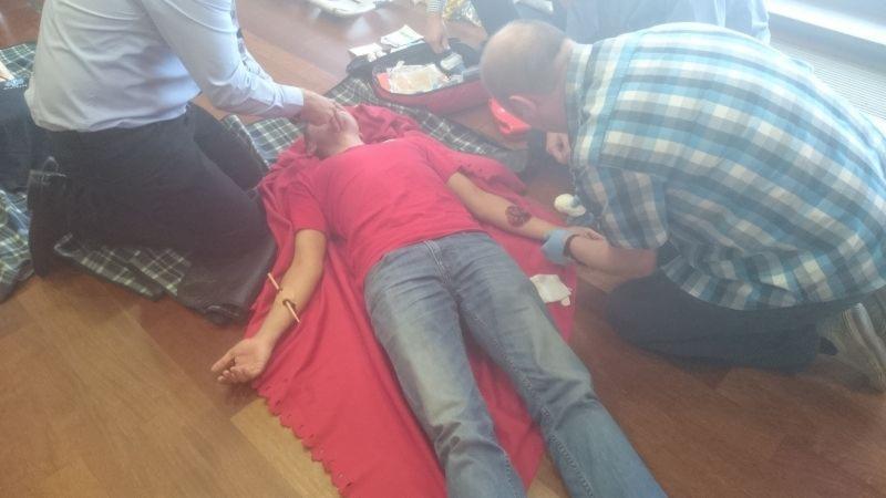 Symulacja udzielania pierwszej pomocy: mężczyzna leży na czerwonym kocu z pozorowanymi ranami na ramionach, opatrywany przez dwie osoby w trakcie szkolenia.