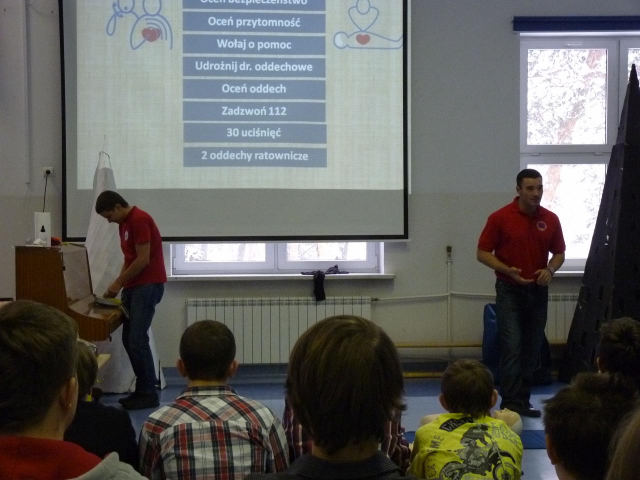 Prelegent w czerwonej koszulce prezentuje zasady udzielania pierwszej pomocy na rzutniku, drugi mężczyzna demonstruje na manekinie, widownia obserwuje.