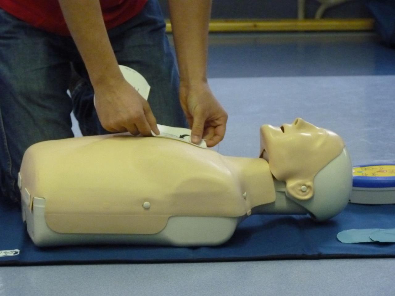 Osoba w dżinsach ćwiczy na manekinie resuscytację, przyklejając elektrody defibrylatora.