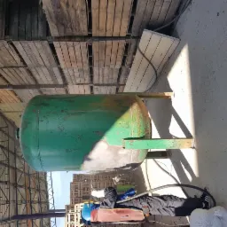 Zielony zbiornik na metalowych nogach, częściowo oczyszczony z rdzy, obok pracownik w kombinezonie ochronnym i masce, widoczne palety w tle.