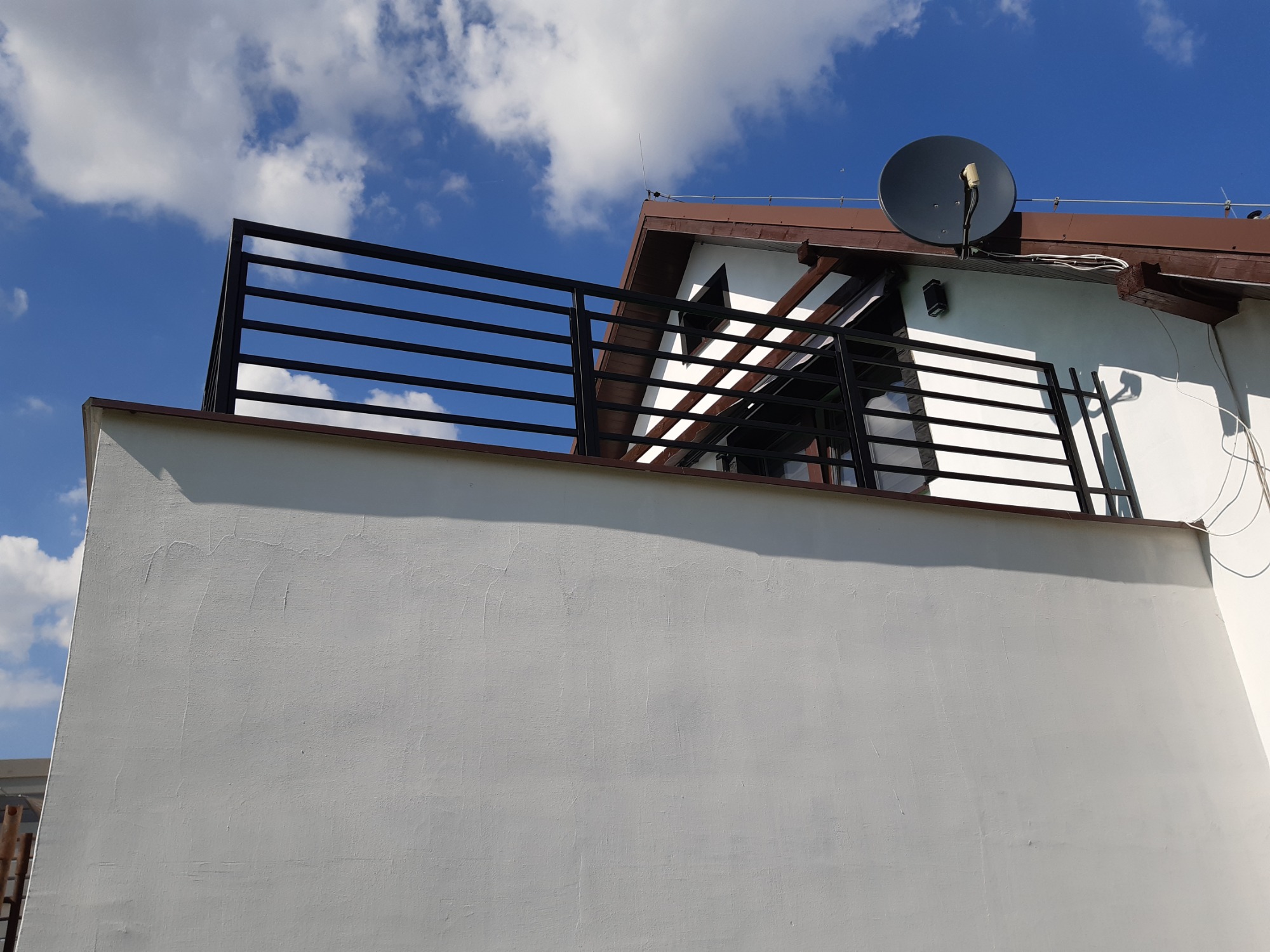 Czarna, pozioma balustrada na balkonie z białymi ścianami, na tle błękitnego nieba z chmurami. Widoczny fragment dachu z anteną satelitarną.