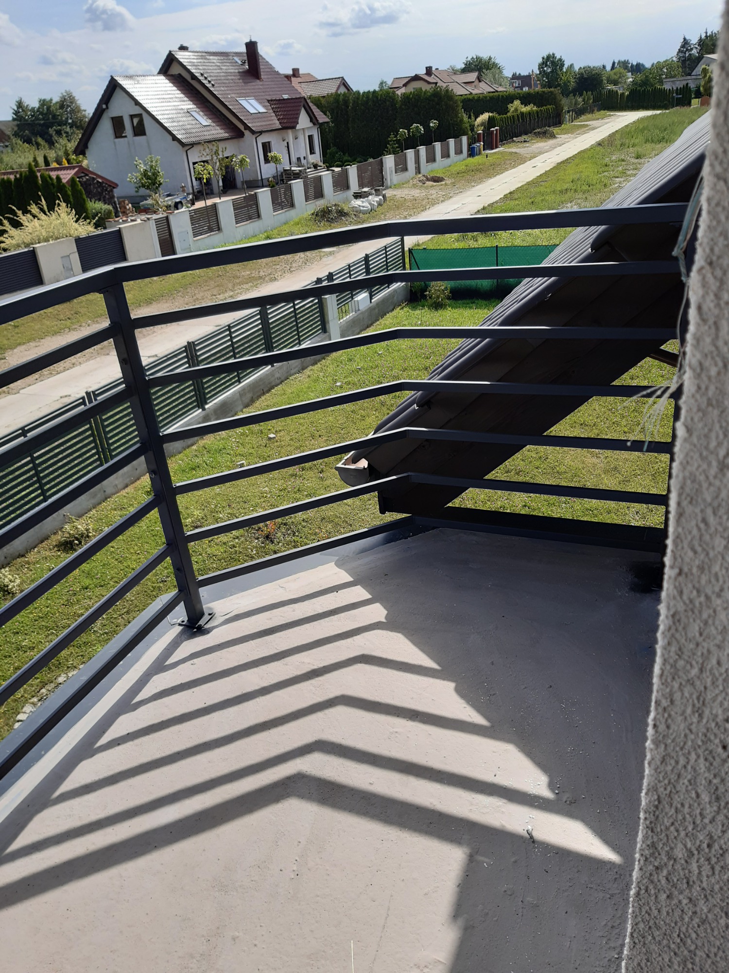 Nowoczesna, czarna balustrada balkonowa z metalowymi poprzeczkami, rzucająca geometryczny cień na betonową podłogę balkonu w Starachowicach, z widokiem na dom i ogrodzenie panelowe w tle.
