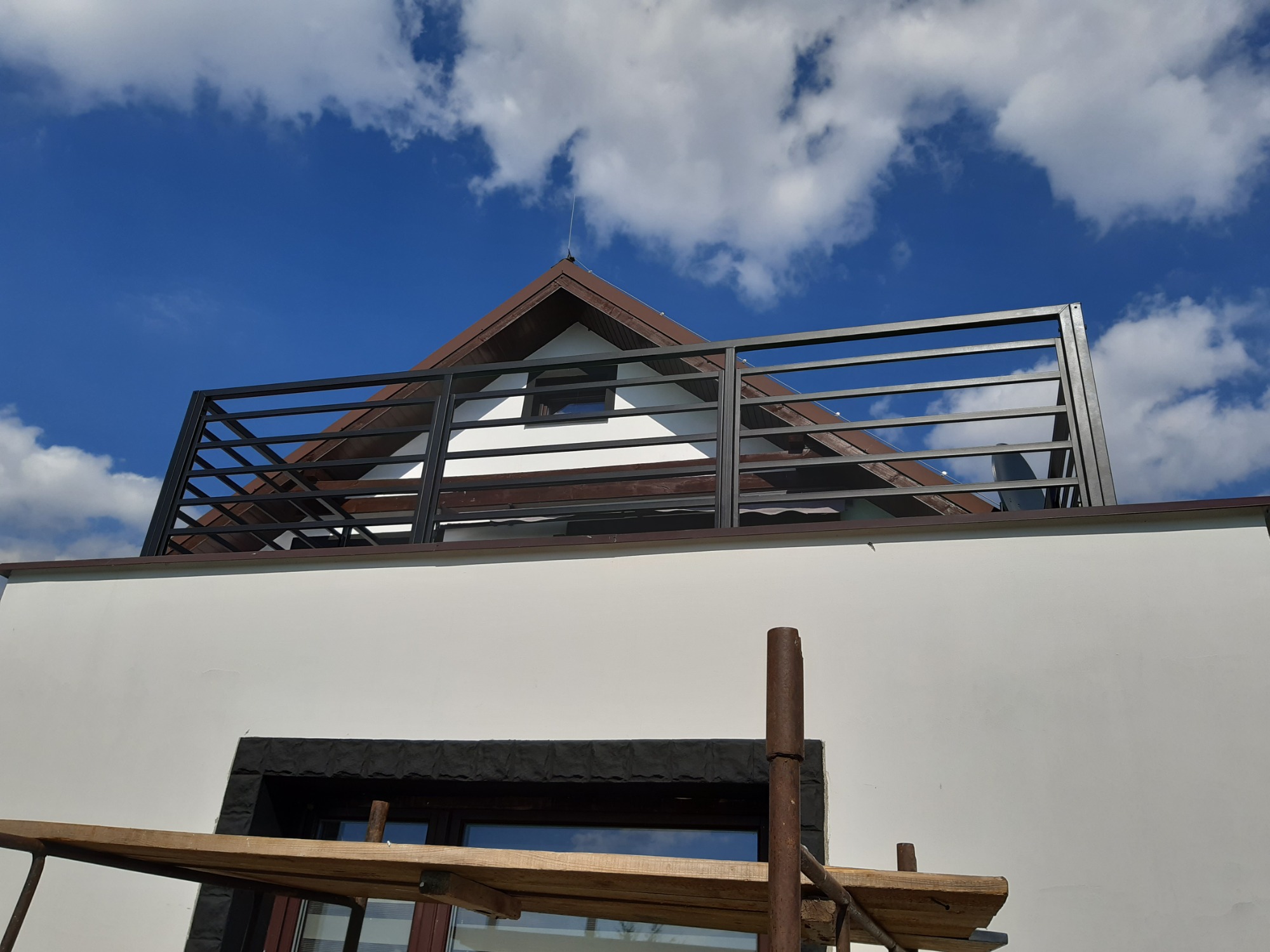 Nowoczesna, czarna balustrada na dachu budynku z białymi ścianami, widoczna z dołu, na tle błękitnego nieba z chmurami, fragment rusztowania w dolnej części kadru.