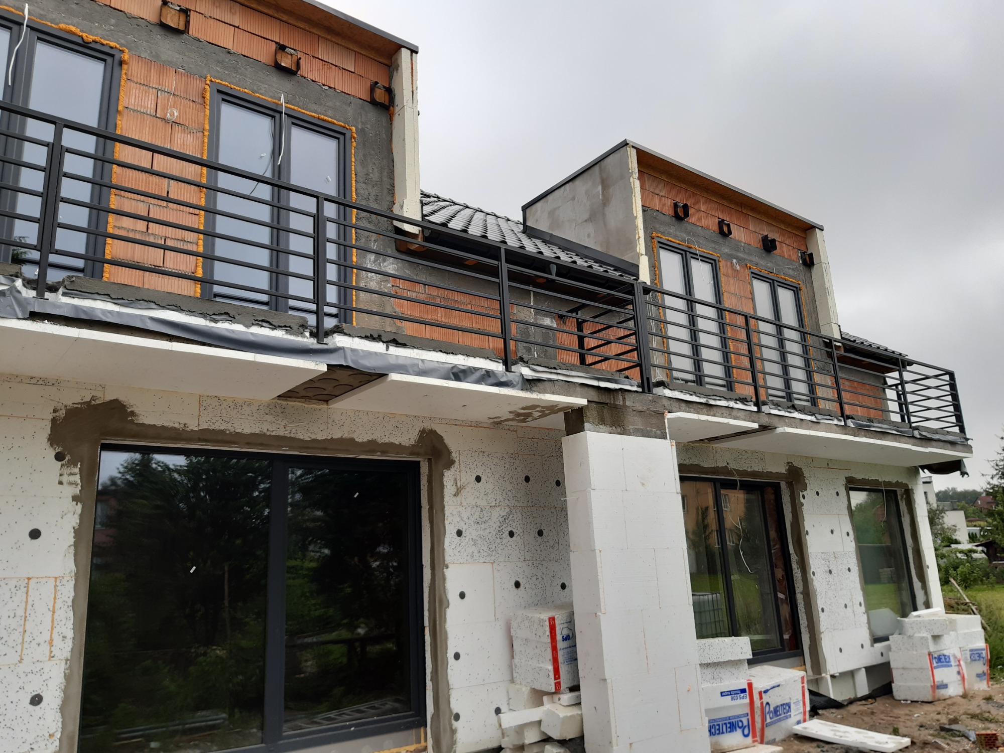 Surowy stan budynku w trakcie ocieplania, z widocznymi czarnymi balustradami balkonowymi i oknami, styropianem elewacyjnym oraz fragmentem dachu z dachówką.