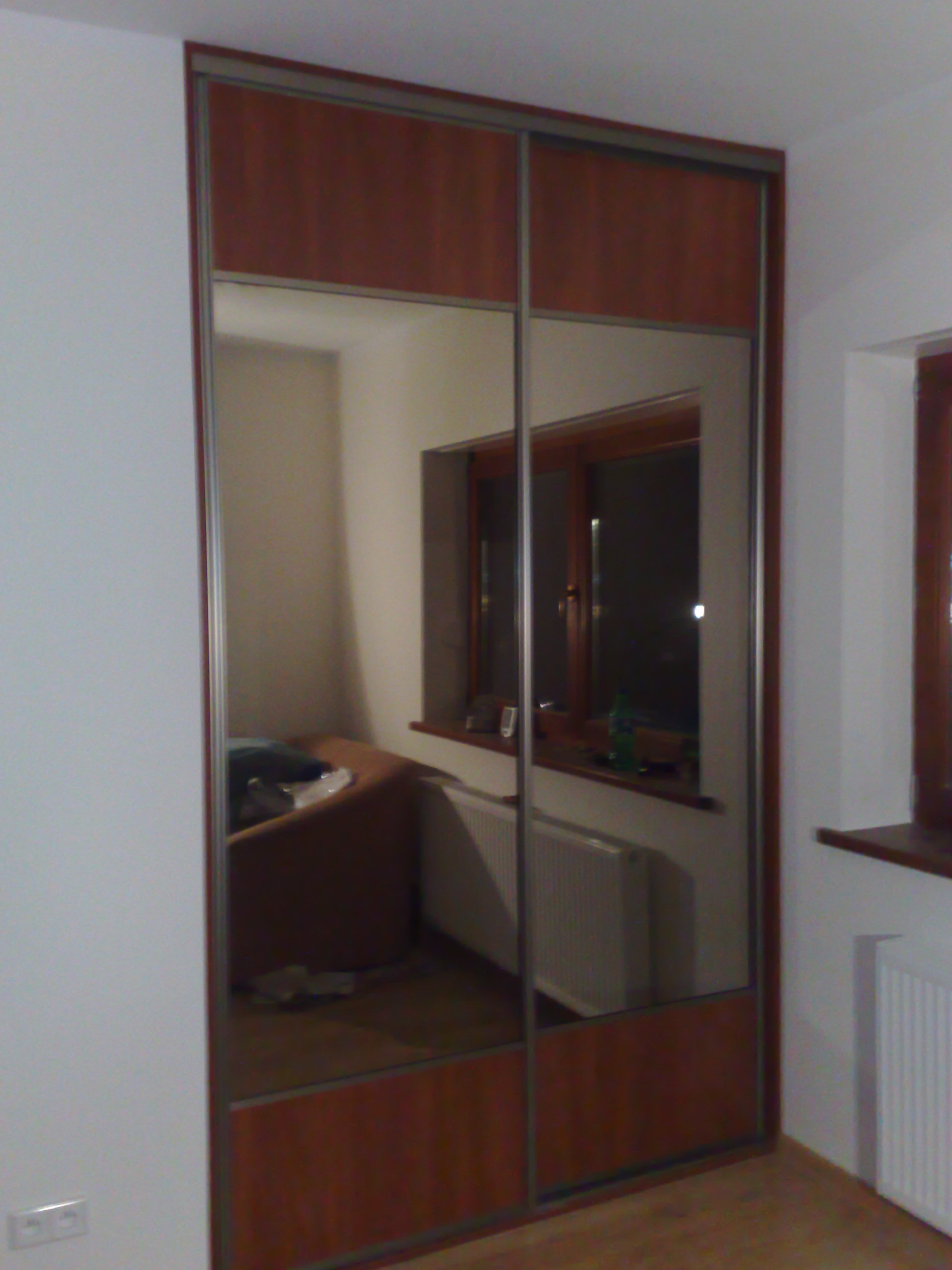 Wnętrze z zabudowaną, dwudrzwiową garderobą z lustrzanymi frontami i drewnianymi panelami w kolorze orzecha, odbijającą pokój z oknem i kaloryferem.