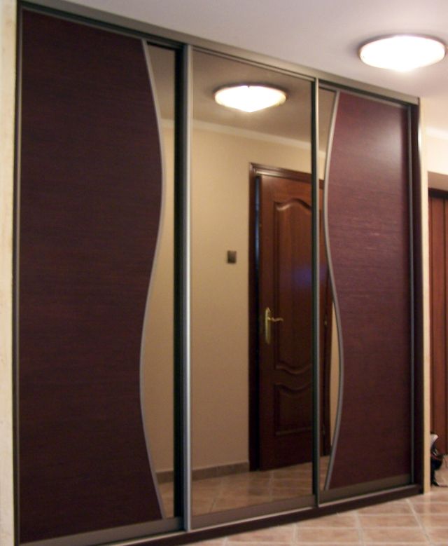 Wnętrze z zabudowaną szafą przesuwną w kolorze ciemnego brązu z lustrzanymi drzwiami, widoczne oświetlenie sufitowe i fragment drzwi w głębi.