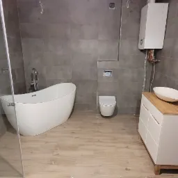 Nowoczesna łazienka z szarymi płytkami, wolnostojącą wanną, szklaną kabiną prysznicową, toaletą podwieszaną i szafką z umywalką nablatową. Widoczne niedokończone wykończenie ściany.