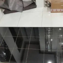 Częściowo wyłożona czarno-biała podłoga z błyszczących płytek, widoczny zwój kabla i elementy instalacji wodnej w ścianie.