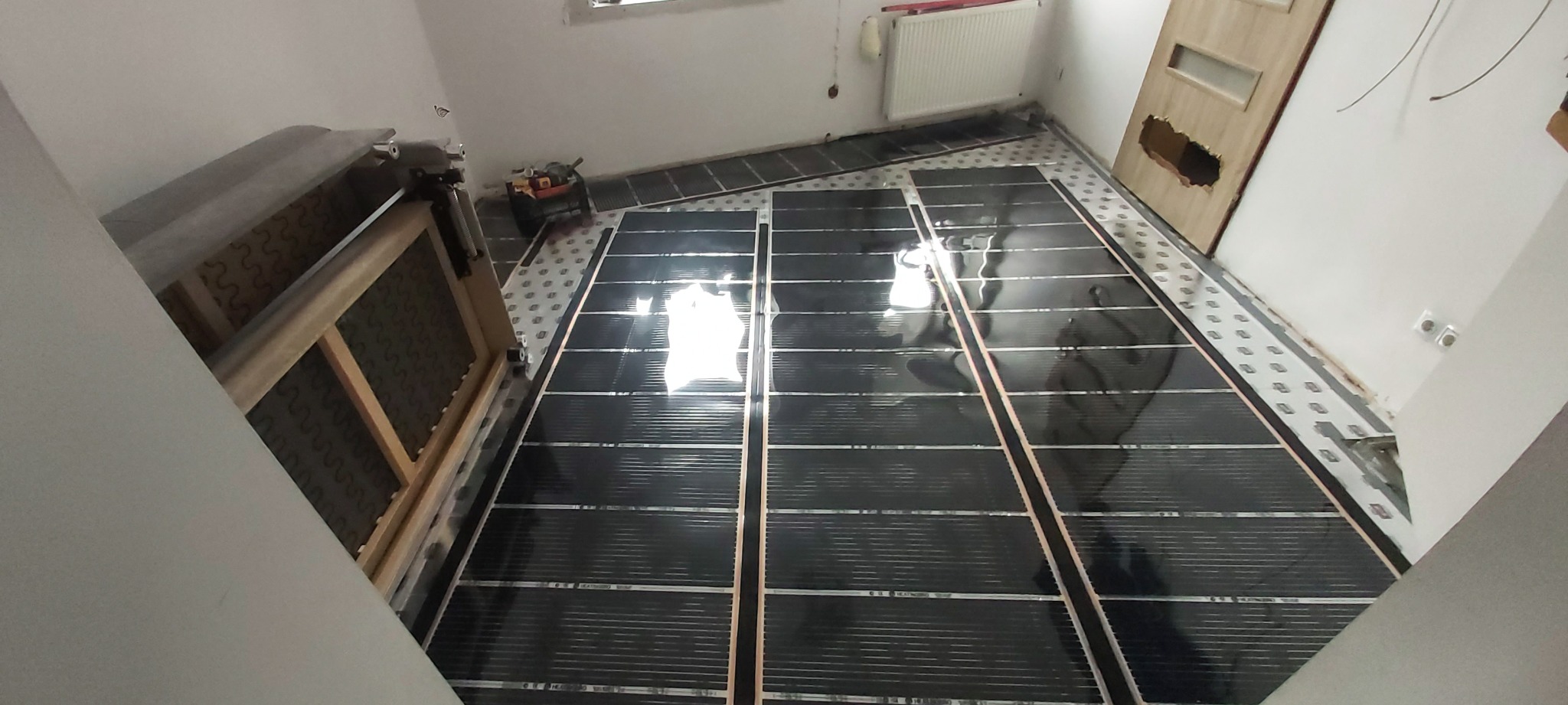 Rozłożone maty grzewcze na podłodze w trakcie montażu, widoczne narzędzia, częściowo zdemontowane drzwi, grzejnik na ścianie