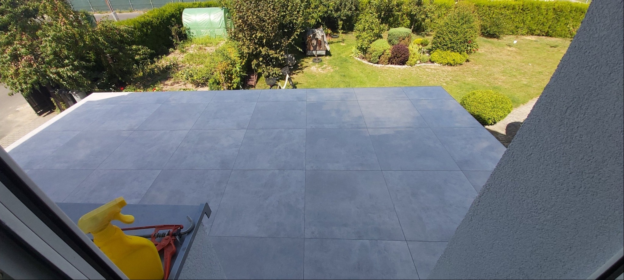 Szare płytki na tarasie, widok z góry, z żółtym spryskiwaczem i narzędziem do cięcia płytek w rogu, z ogrodem w tle.