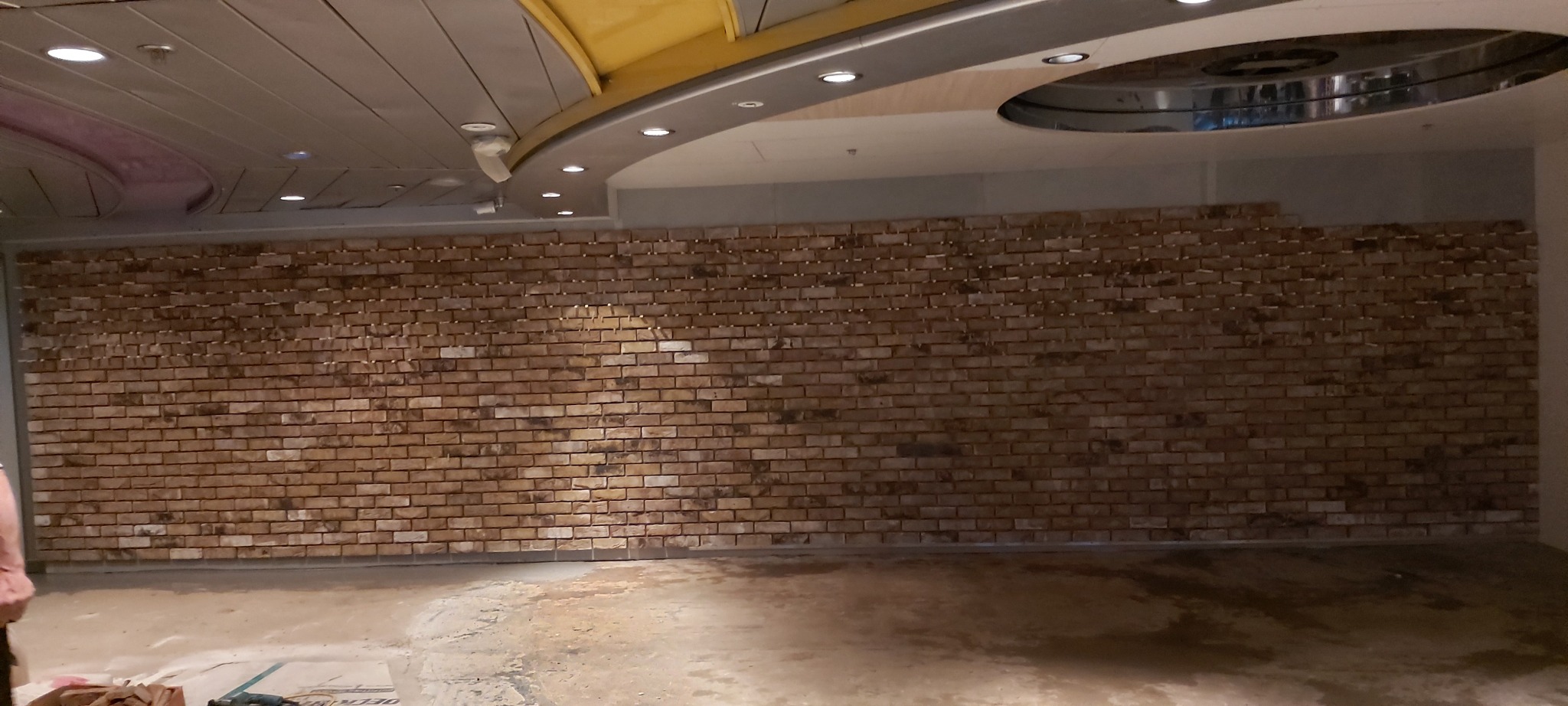 Ściana w trakcie wykańczania z cegieł klinkierowych w pomieszczeniu z surowym wykończeniem podłogi i sufitu, widoczne narzędzia glazurnicze.