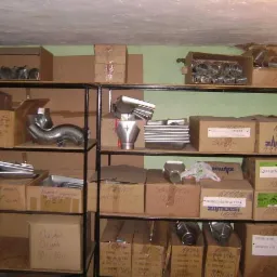 Zdjęcie przedstawia magazynowane elementy metalowe, prawdopodobnie rynny, ułożone na metalowych regałach w kartonowych pudełkach z widocznymi etykietami i napisami, co sugeruje stan przed montażem.