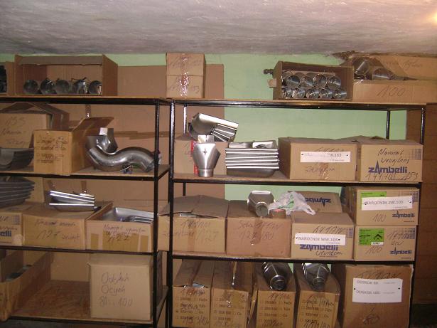 Zdjęcie przedstawia magazynowane elementy metalowe, prawdopodobnie rynny, ułożone na metalowych regałach w kartonowych pudełkach z widocznymi etykietami i napisami, co sugeruje stan przed montażem.