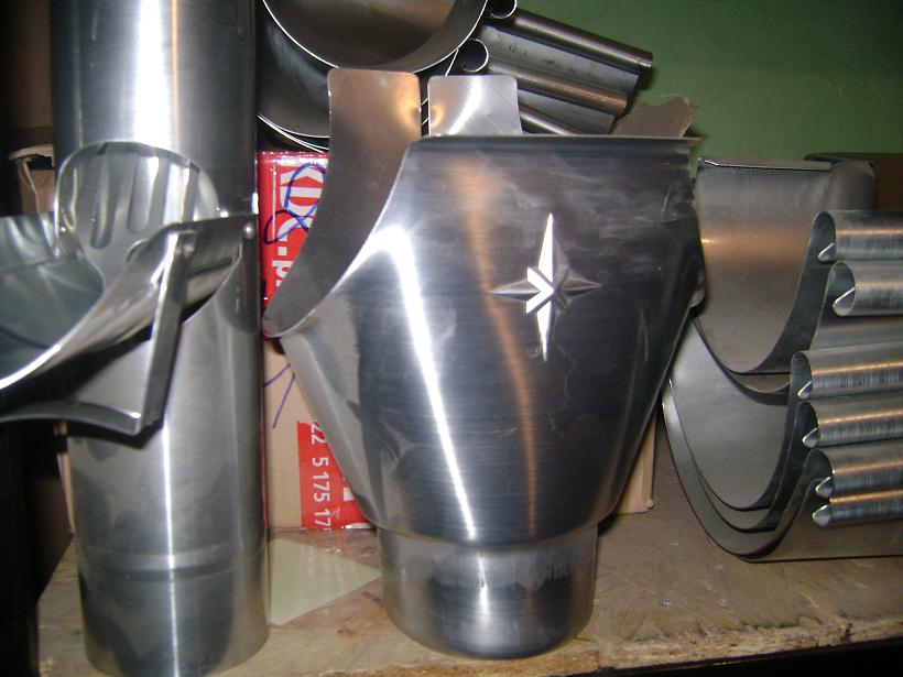 Stalowe elementy systemu odprowadzania wody deszczowej, w tym kształtki i rury spustowe, z widocznym wytłoczonym znakiem w kształcie gwiazdy na jednym z elementów.