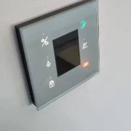 Szary panel dotykowy Ampio zamontowany na ścianie, z ikonami sterowania oświetleniem, temperaturą, roletami i zamkiem, niektóre podświetlone na zielono i czerwono.