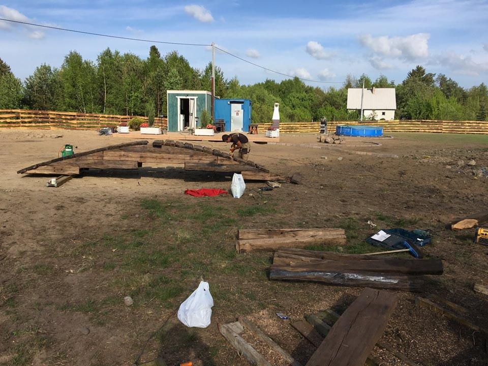 Budowa drewnianego placu zabaw z elementami konstrukcyjnymi na ziemi, widoczny pracownik w trakcie montażu, w tle budynki gospodarcze, ogrodzenie i basen ogrodowy.