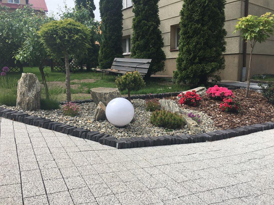 Ogród z dekoracyjną kulą i kamieniami, obrzeża z ciemnej kostki, tło z iglakami i ławką, widok z poziomu brukowanego chodnika.