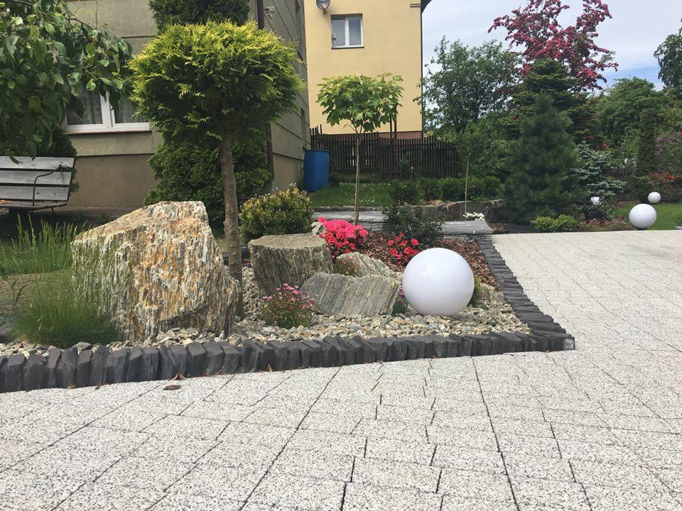 Ogród z dekoracyjnymi kamieniami, kulami świetlnymi i równo ułożoną kostką brukową w odcieniach szarości, obrzeża z ciemnego kamienia.