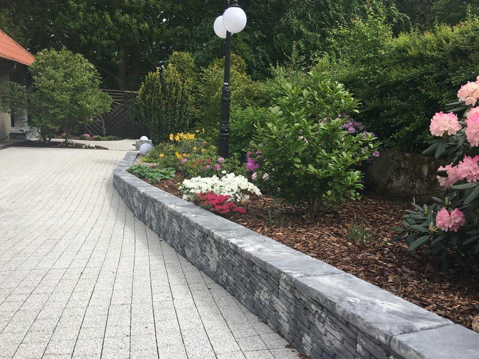 Ogród z podwyższoną rabatą kwiatową obłożoną kamieniem łupanym, oświetloną lampą parkową, z kostką brukową na pierwszym planie i różanecznikami w tle.