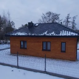 Drewniany dom jednorodzinny z grafitowym dachem pokrytym częściowo śniegiem, widziany zza metalowego ogrodzenia w zimowej scenerii.