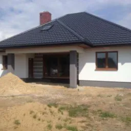 Dom jednorodzinny z białą elewacją, ciemnym dachem i szarymi kolumnami podtrzymującymi zadaszenie tarasu. Na pierwszym planie hałda piasku.