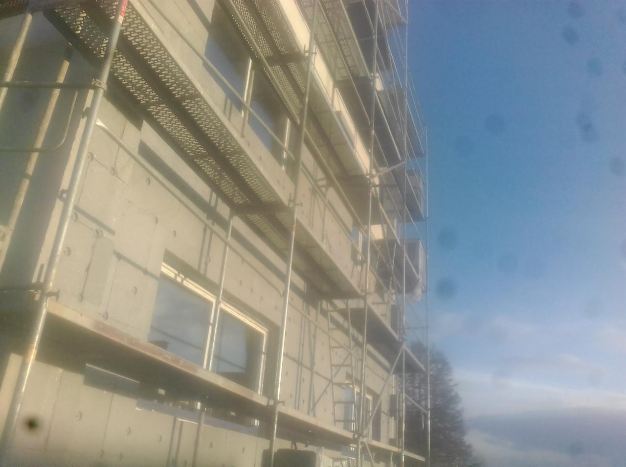 Budynek w trakcie ocieplania, widoczne rusztowania i płyty izolacyjne na elewacji, fragment niebieskiego nieba w tle.