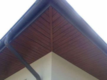 Fragment narożnika dachu z brązową podbitką drewnianą ułożoną w jodełkę i czarną rynną spustową na tle jasnej ściany budynku.