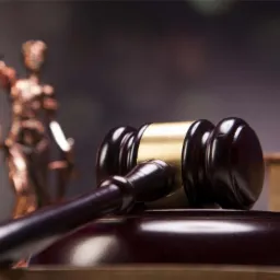 Drewniany młotek sędziowski z pozłacanym elementem, na podstawie, obok statuetki Temidy z wagą sprawiedliwości w tle.