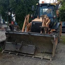 Żółto-czarna koparko-ładowarka CASE 580 SR na żwirowym podłożu, widoczne ślady opon, w tle drzewa i budynek.