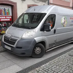 Srebrny bus Citroen zaparkowany na chodniku z kostki, obok sklepu z chemią i zabawkami, z logo firmy transportowej na boku, oferującej usługi transportowe i przeprowadzki.