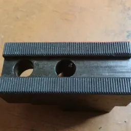 Szczegółowe ujęcie czarnego, metalowego uchwytu tokarskiego z dwoma otworami i rowkowaną powierzchnią, leżącego na drewnianym stole.