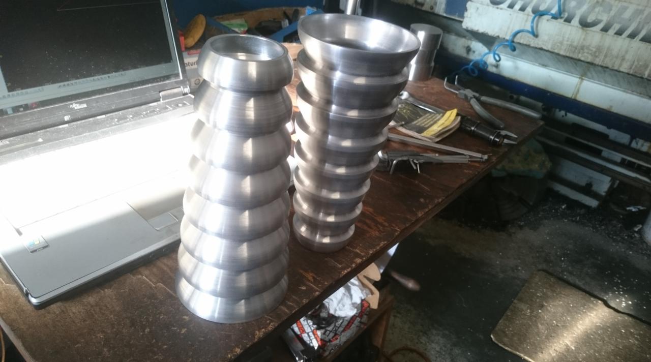 Dwa metalowe elementy obrobione na tokarce, leżące na warsztatowym stole obok laptopa i narzędzi pomiarowych, w tle widoczna maszyna.