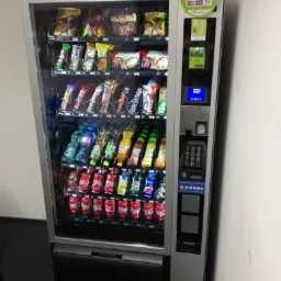 Automat vendingowy wypełniony przekąskami, napojami gazowanymi i wodą butelkowaną, z panelem płatności zbliżeniowych i klawiaturą numeryczną.