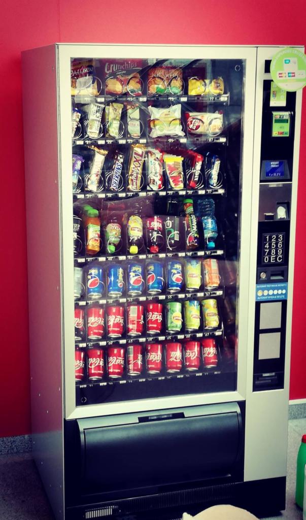 Automat vendingowy z napojami i przekąskami, widoczne Coca-Cola, Pepsi, Snickers i Crunchips na tle czerwonej ściany.