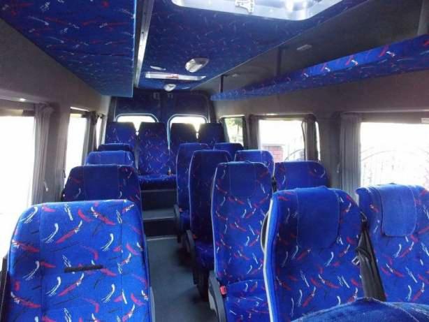 Wnętrze autobusu z niebieskimi fotelami w abstrakcyjne wzory, widok z przodu kabiny pasażerskiej, oświetlenie sufitowe.