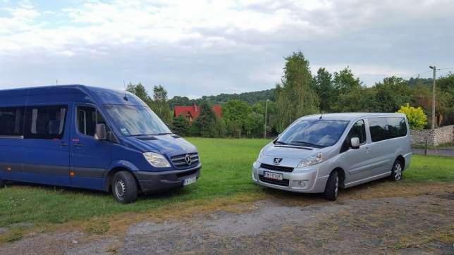 Dwa busy, niebieski Mercedes i srebrny Peugeot, stoją na trawiastym terenie z zielonymi drzewami w tle.