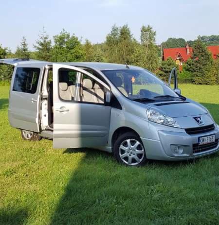 Srebrny van Peugeot z otwartymi drzwiami bocznymi i tylnymi, zaparkowany na zielonej trawie przed domem i drzewami.