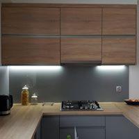 Nowoczesna kuchnia: górne szafki z frontami imitującymi drewno, dolne szafki w kolorze szarym, szary panel nad blatem, oświetlenie LED podszafkowe, płyta gazowa.