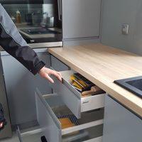 Szare szafki kuchenne z drewnianym blatem; otwarta szuflada z organizerem na przybory kuchenne; widoczna dłoń mężczyzny.