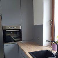 Szara zabudowa kuchenna z piekarnikiem w stali nierdzewnej, jasny drewniany blat, czarny zlew i bateria przy oknie.