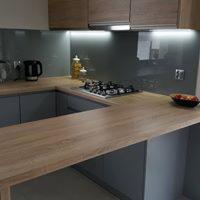 Szare szafki kuchenne z drewnianym blatem i szklanym panelem nad blatem, oświetlenie LED pod szafkami wiszącymi, czajnik i przyprawy na blacie.