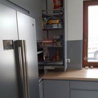 Szara kuchnia z wysuwanym koszem cargo obok lodówki, blat drewnopodobny, okno w tle.