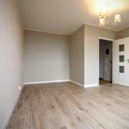 Pomalowane na beżowo puste pomieszczenie z nową podłogą z paneli, otwartymi białymi drzwiami i ozdobnym żyrandolem.